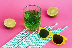 sunglasses and glass of green alcohol drink near striped paper straws and limes on pink