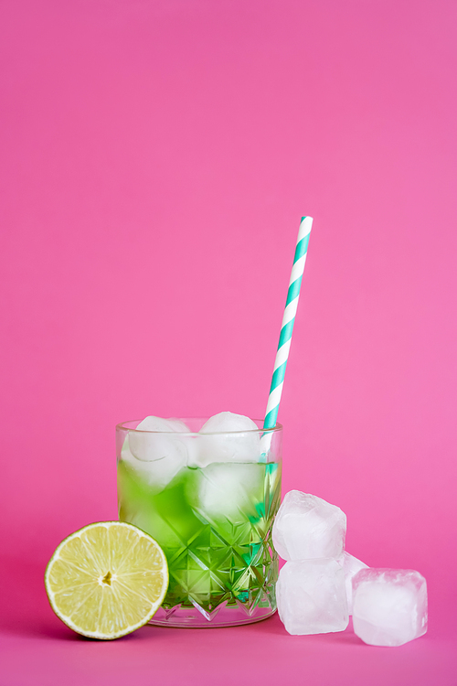 frozen ice cubes in glass with green mojito drink with straw near lime on pink