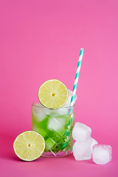 frozen ice cubes in glass with green mojito drink with straw and limes on pink