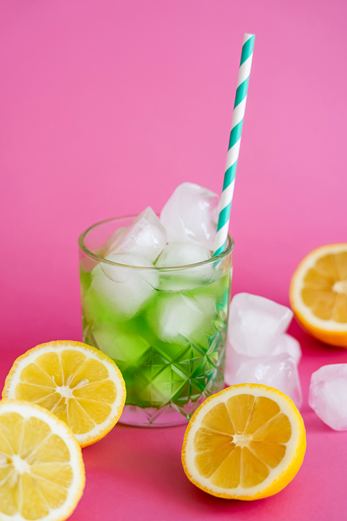 frozen ice cubes in glass with green mojito drink and straw near lemons on pink