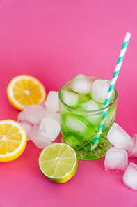 ice cubes in glass with mojito drink and straw near citrus fruits on pink