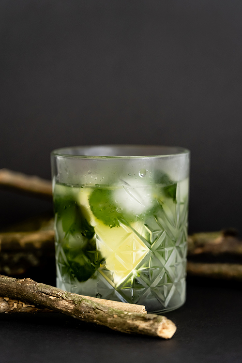 faceted cold glass with melting ice cubes and sliced lime near wooden sticks on black
