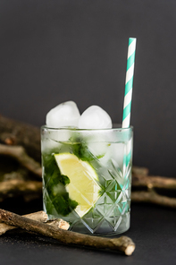 faceted cold glass with ice cubes and sliced lime near wooden sticks on black