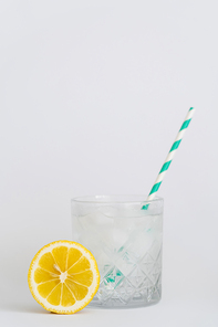 faceted cold glass with ice cubes and paper straw near sliced lemon on white