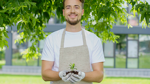 Smiling gardener in gloves holding plant in soil in garden