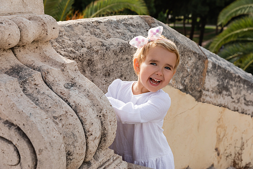 Cheerful baby girl in dress looking at camera near stone Puente Del Mar bridge in Valencia
