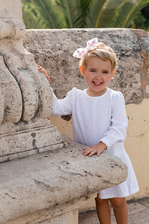 Positive toddler child in summer dress looking at camera near stone Puente Del Mar bridge in Valencia