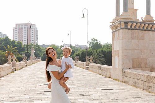 Happy woman holding baby in white dress and looking at camera on Puente Del Mar bridge in Valencia