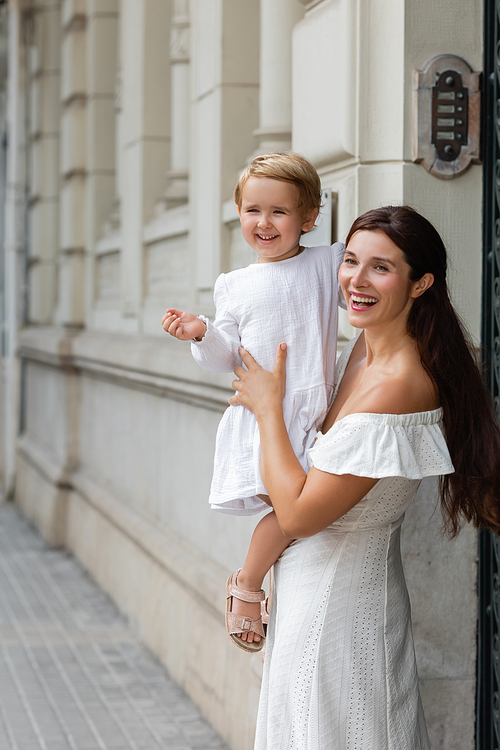 Smiling brunette woman in summer dress holding toddler girl near building in Valencia