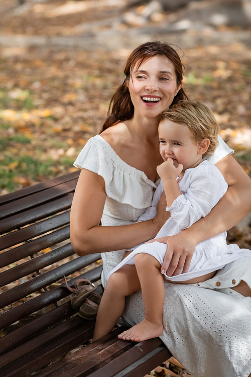 Brunette woman hugging toddler girl in summer dress on bench in park