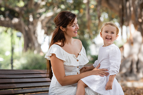Brunette woman looking at daughter laughing on bench in park