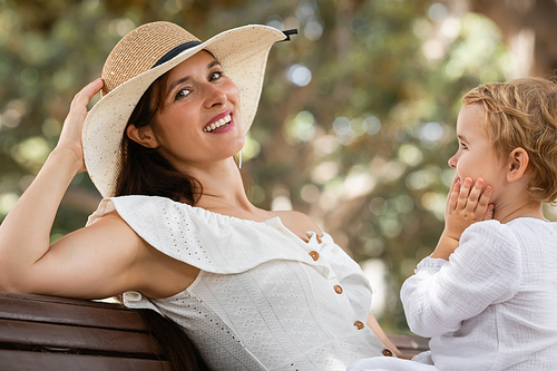 Smiling woman in sun hat and dress looking at camera near child on bench in park