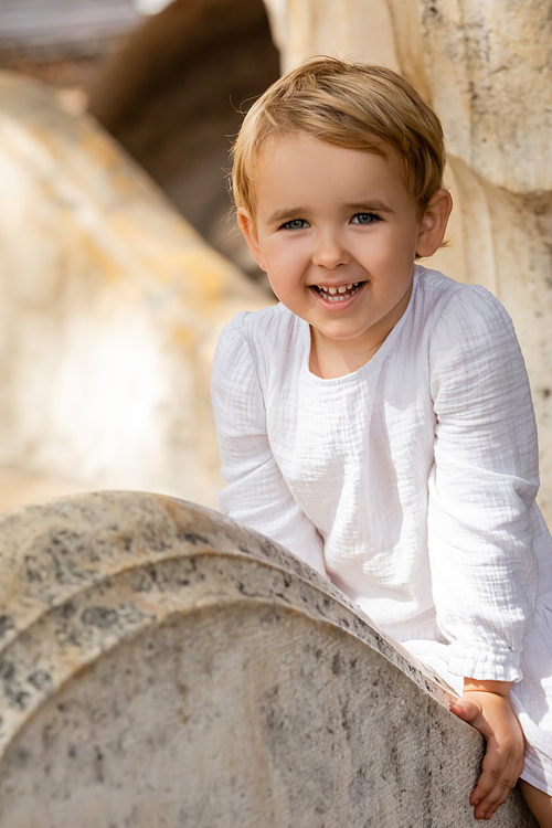 Smiling baby girl in white dress sitting on stone bench in park