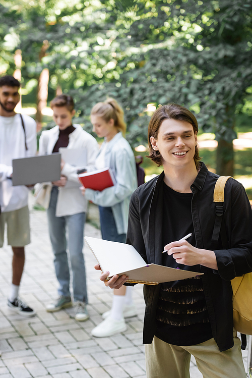 Smiling student holding notebook near blurred interracial friends in park