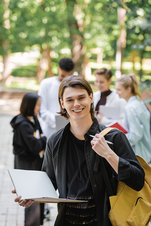 Smiling student with notebook and backpack looking at camera near blurred friends in park