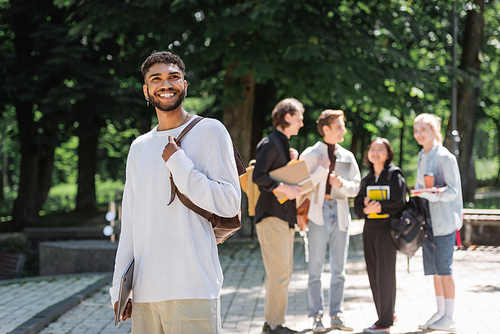 Smiling african american student with laptop standing near blurred friends in summer park