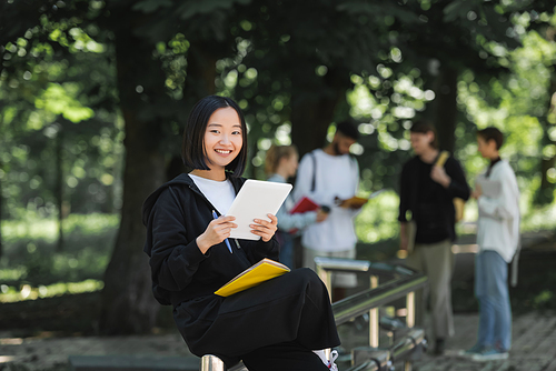 Cheerful asian student with digital tablet and notebook looking at camera in park