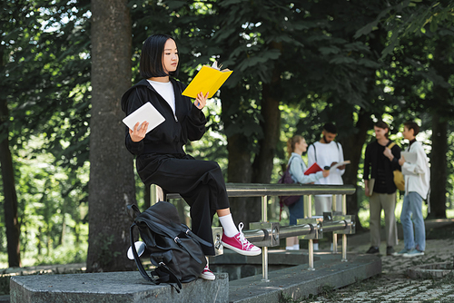 Asian student reading book and holding digital tablet in park
