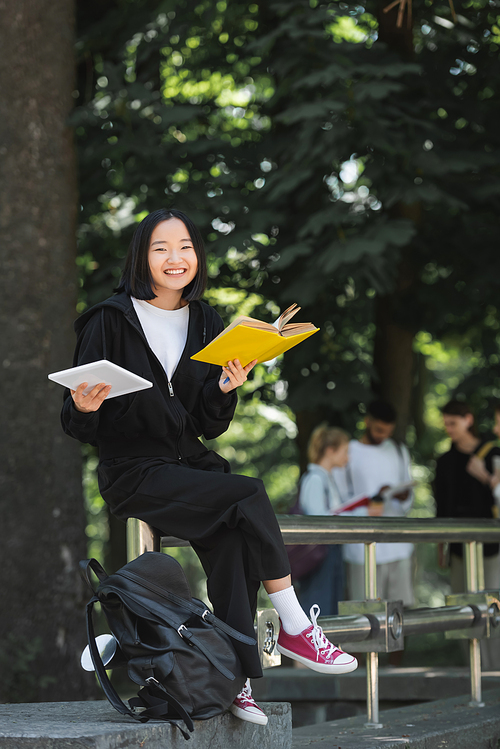Cheerful asian student with book and digital tablet looking at camera near backpack in park