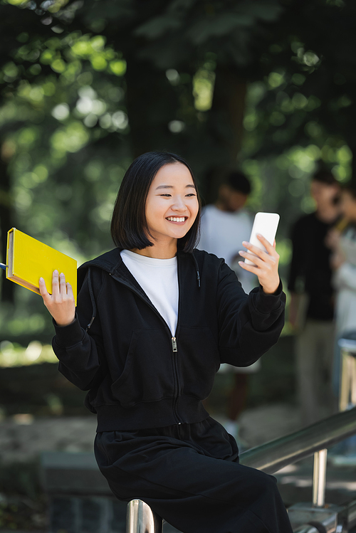 Smiling asian student holding book and taking selfie on smartphone in park