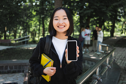 Cheerful asian student with book showing smartphone in park