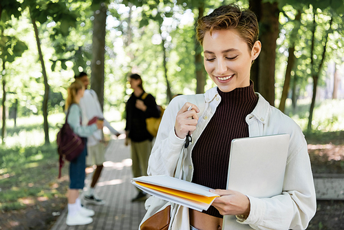Cheerful student with laptop looking at notebook in summer park