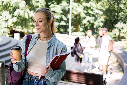 Smiling student holding coffee to go and notebook in park