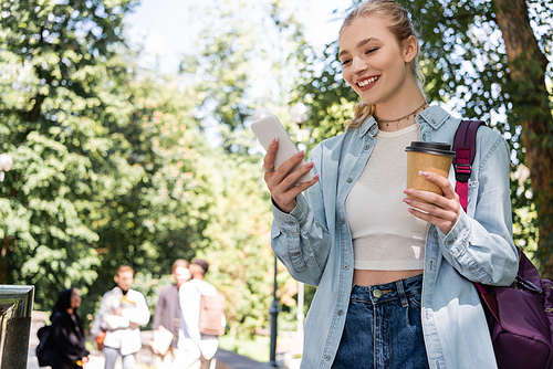 Smiling student using smartphone and holding coffee to go in park