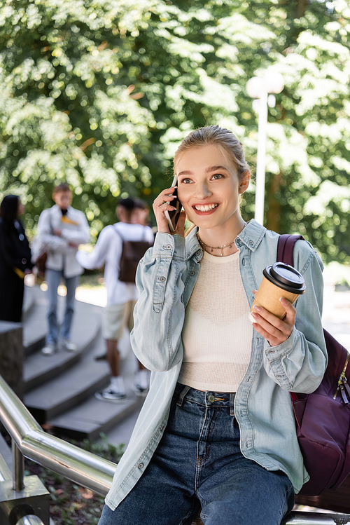 Smiling student with coffee to go and backpack talking on smartphone in park