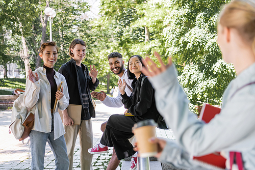 Smiling multiethnic students waving hands at blurred friend in park