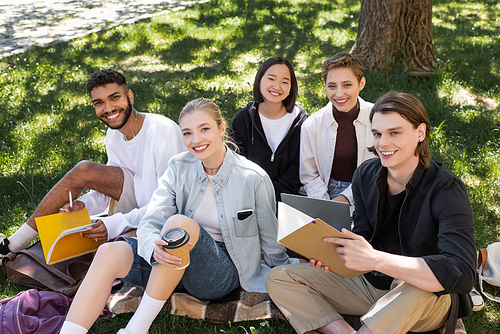 Smiling interracial students holding notebooks and coffee to go on grass in park