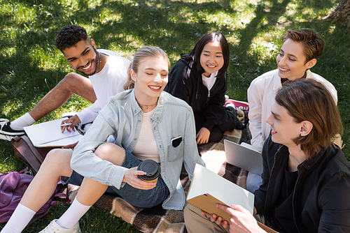 High angle view of cheerful multicultural students with copy books and devices sitting on grass in park