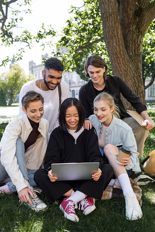 Cheerful interracial students looking at laptop on grass in park