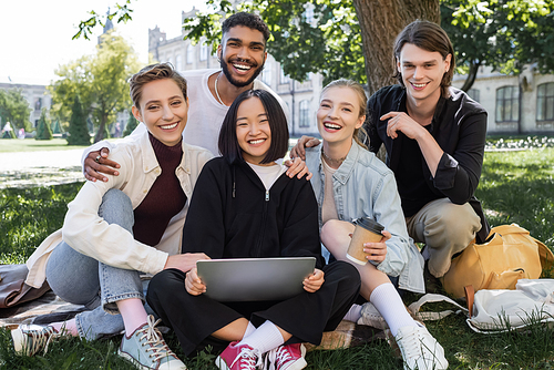 Positive multicultural students with laptop hugging on grass in park