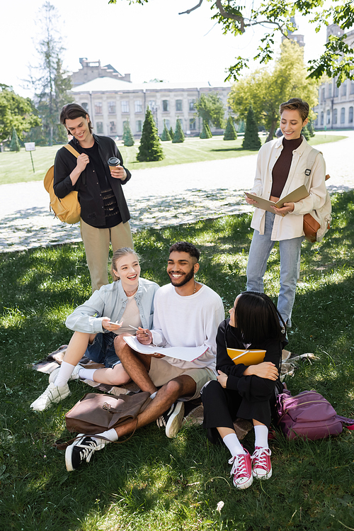 Cheerful interracial students with copy books spending time on grass in park