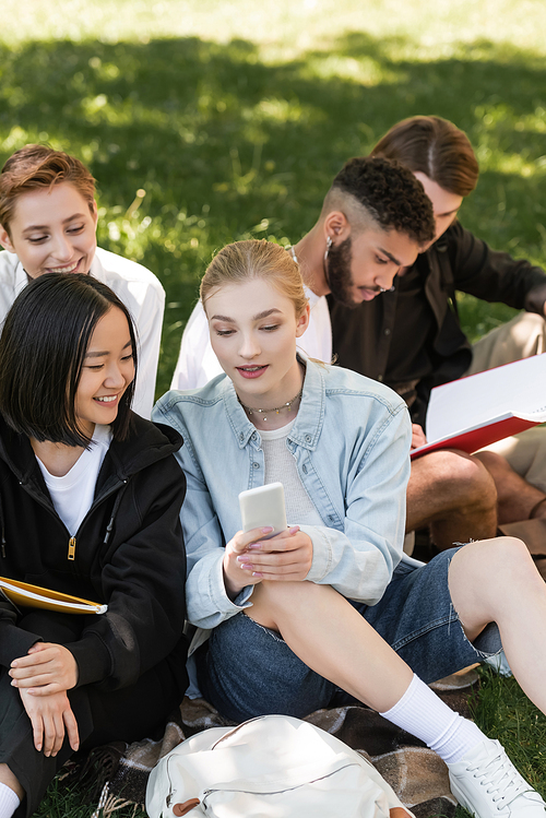 Student holding smartphone near multicultural friends on lawn in summer park