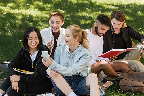 Positive student holding smartphone near multiethnic friends on grass outdoors
