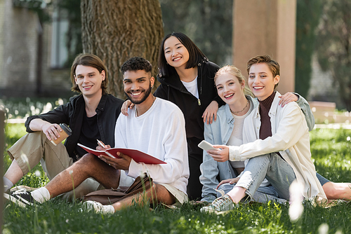 Cheerful interracial students with cellphone and copy book looking at camera in park