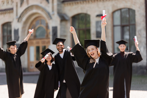 Excited student in bachelor gown holding diploma near multiethnic friends outdoors