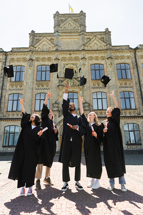 Positive interracial bachelors with diplomas throwing caps near university outdoors