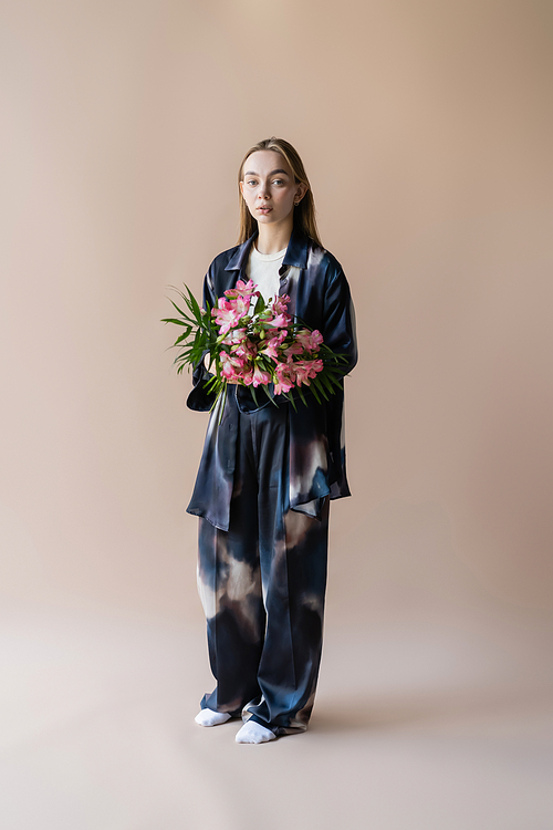full length of woman in tie-dye style clothes standing with floral bouquet on beige background