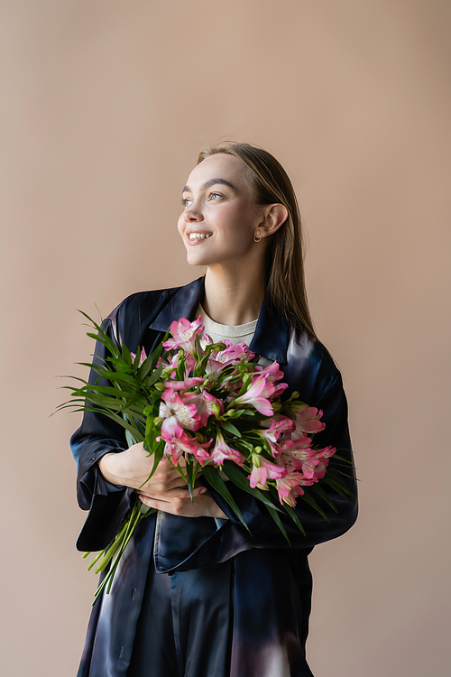 happy and trendy woman with fresh floral bouquet looking away isolated on beige