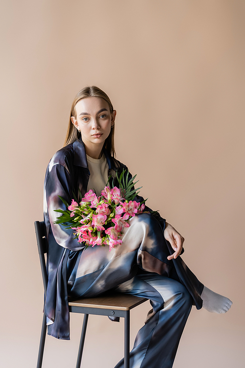 young woman in gradient clothes sitting on chair with alstroemeria flowers isolated on beige