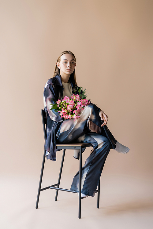 full length of stylish woman with alstroemeria bouquet posing on chair on beige background