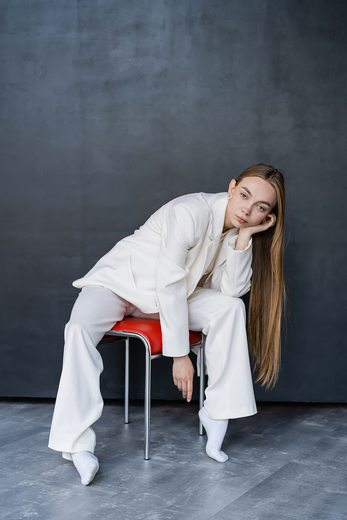 full length of stylish woman in white suit and socks sitting on chair on black background