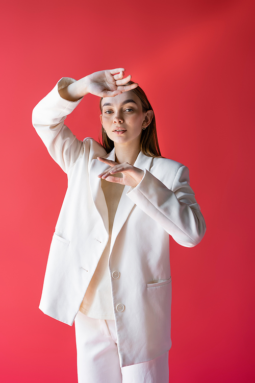 young and stylish woman in white blazer looking at camera while posing isolated on pink