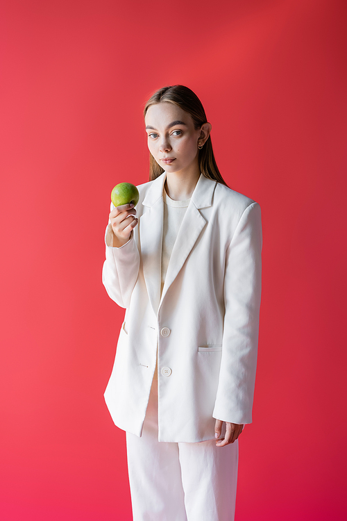 pretty woman in white blazer holding fresh apple and looking at camera isolated on crimson