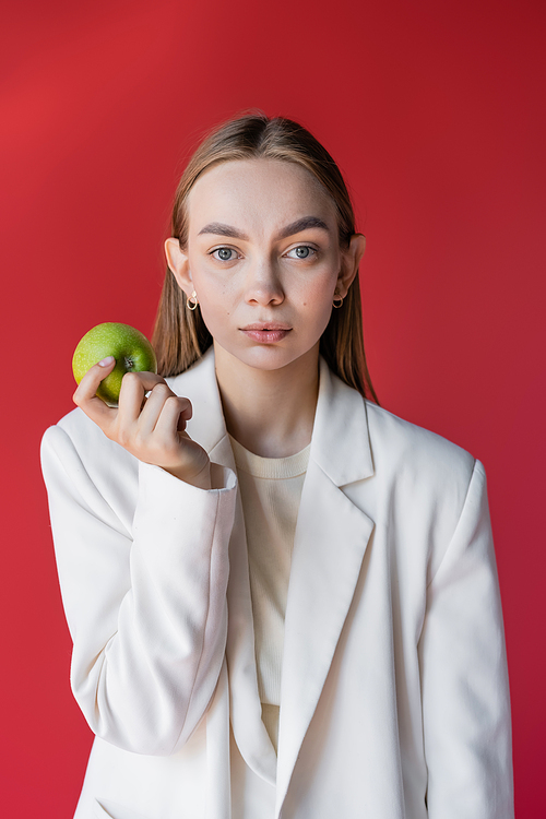 fashionable woman in white blazer holding ripe apple isolated on crimson