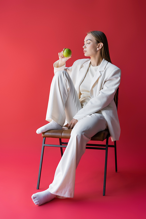 full length of woman in white suit and socks sitting on chair with apple on pink background