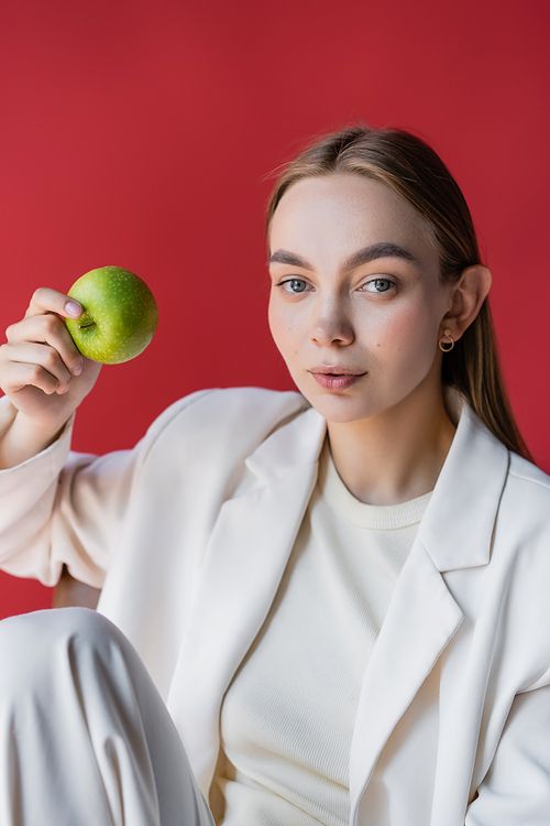 pretty and trendy woman with juicy apple looking at camera isolated on crimson